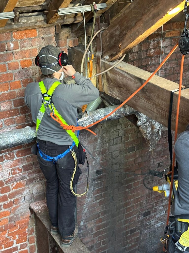 A man strapped to ropes connected to roof beams, standing on a thin wooden platform investigates the join between brickwork and the beams.